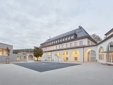 Schulgebäude mit Arkadengang und großem gepflastertem Schulhof bei bewölktem Himmel