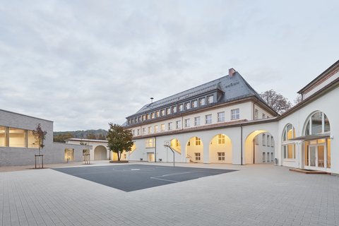 Schulgebäude mit Arkadengang und großem gepflastertem Schulhof bei bewölktem Himmel