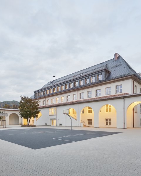 Schulgebäude mit Arkadengang und großem gepflastertem Schulhof bei bewölktem Himmel