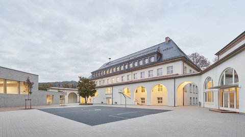 Schulgebäude mit Arkadengang und großem gepflastertem Schulhof bei bewölktem Himmel