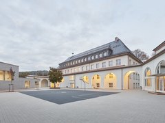 Schulgebäude mit Arkadengang und großem gepflastertem Schulhof bei bewölktem Himmel