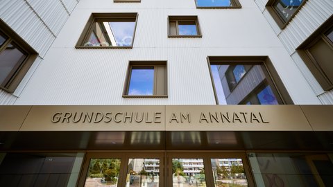 Blick von unten auf die Fassade eines mehrstöckigen Gebäudes mit rechteckigen Fenstern und dem Schriftzug 'GRUNDSCHULE AM ANNATAL' über dem Eingang