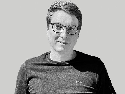 Schwarzweiß-bild eines lächelnden Mannes mit kurzem Haar und runder Brille, bekleidet mit einem dunklen T‑Shirt, vor hellem einfarbigem Hintergrund; entspannte, leicht zur Kamera gedrehte Haltung.