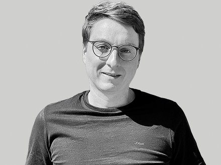 Schwarzweiß-bild eines lächelnden Mannes mit kurzem Haar und runder Brille, bekleidet mit einem dunklen T‑Shirt, vor hellem einfarbigem Hintergrund; entspannte, leicht zur Kamera gedrehte Haltung.