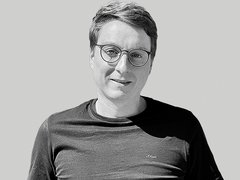 Schwarzweiß-bild eines lächelnden Mannes mit kurzem Haar und runder Brille, bekleidet mit einem dunklen T‑Shirt, vor hellem einfarbigem Hintergrund; entspannte, leicht zur Kamera gedrehte Haltung.