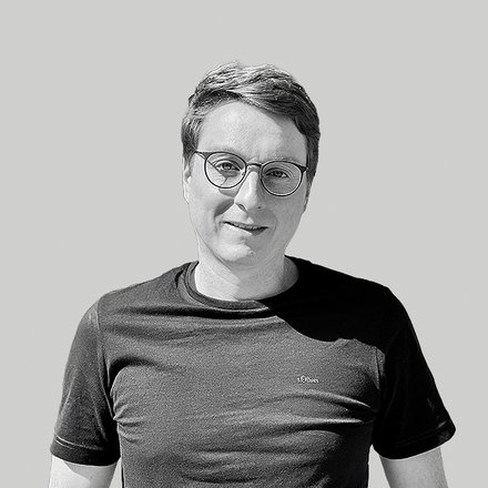 Schwarzweiß-bild eines lächelnden Mannes mit kurzem Haar und runder Brille, bekleidet mit einem dunklen T‑Shirt, vor hellem einfarbigem Hintergrund; entspannte, leicht zur Kamera gedrehte Haltung.