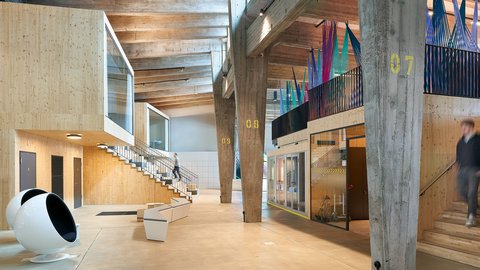 "Heller Innenraum mit rohem Holz und Betonkonstruktion. Mehrere Ebenen verbunden durch Treppen, Leitern und Rutschen in verschiedenen Farben. Ein großes weißes Spielgerät links, Beschilderung mit Nummern. Personen in Bewegung auf den Treppen und Galerien."