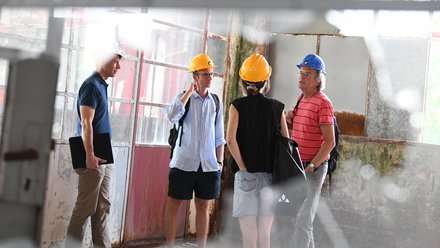 Vier Personen mit Schutzhelmen stehen in einer alten, teils verrosteten Industriehalle und besprechen die Gebäudebegutachtung.