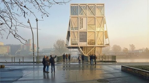 "Modernes Kulturhaus mit großer Glasfassade und Holzrahmen-Struktur steht auf einer feuchten Plattform an einem Fluss. Mehrere Besucher stehen davor. Im Hintergrund sind Industriegebäude und kahle Bäume zu sehen. Der Himmel ist bewölkt."