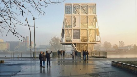 "Modernes Kulturhaus mit großer Glasfassade und Holzrahmen-Struktur steht auf einer feuchten Plattform an einem Fluss. Mehrere Besucher stehen davor. Im Hintergrund sind Industriegebäude und kahle Bäume zu sehen. Der Himmel ist bewölkt."