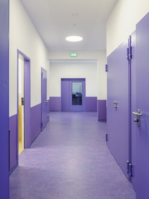 Ein heller, moderner Flur im Innenraum. Der Boden ist in einem kräftigen Lilaton gehalten, die Wände sind weiß mit einem lila Sockel. Mehrere Türen, darunter eine mit Glasausschnitt am Ende des Flurs, sind ebenfalls lila. Eine runde Deckenleuchte erhellt den Gang.