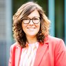 Porträt einer Person: Eine Person mit lockigem Haar trägt eine rosafarbene Jacke und ein gemustertes Oberteil und lächelt in die Kamera.