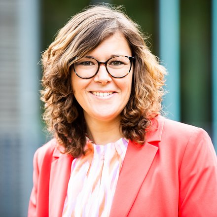 Porträt einer Person: Eine Person mit lockigem Haar trägt eine rosafarbene Jacke und ein gemustertes Oberteil und lächelt in die Kamera.