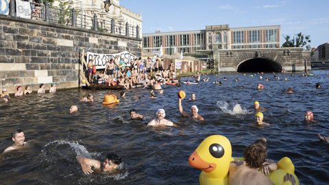 Viele Menschen schwimmen an einem Flussufer vor einer historischen Ufermauer. Weitere Personen sitzen auf einem Steg oder beobachten das Geschehen vom Ufer aus.
