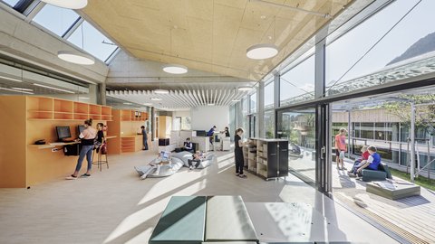 Heller, offener Bibliotheks- und Aufenthaltsbereich mit orangefarbenen Regalen, Computern, Sitzsäcken und niedrigen Bücherregalen. Schülerinnen und Schüler lernen, lesen oder sitzen im Gespräch; rechts öffnet sich der Raum zu einer Terrasse.