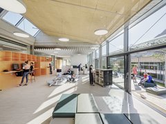 Heller, offener Bibliotheks- und Aufenthaltsbereich mit orangefarbenen Regalen, Computern, Sitzsäcken und niedrigen Bücherregalen. Schülerinnen und Schüler lernen, lesen oder sitzen im Gespräch; rechts öffnet sich der Raum zu einer Terrasse.