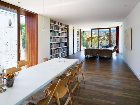 "Moderner, offener Wohnbereich mit großzügiger Architektur. Im Vordergrund ein langer weißer Esstisch mit sechs hölzernen Stühlen im Vintage-Design. Am Tisch links ein Glasvase mit gelben Blüten und ein Kerzenständer. Dahinter ein großes Bücherregal mit dicht besetzten Regalfächern. Die linke Wand wird von bodenhohen Terrassentüren mit rotem Holzrahmen dominiert, dahinter grüne Vegetation sichtbar. Rechts weitere Fenster mit Blick auf ein blaues Meer. Im Hintergrund ein Arbeitsbereich mit Schreibtisch. Der Boden ist mit dunklem Holzparkett belegt. Moderne Hängeleuchten mit grauer Färbung hängen von der weißen Decke herab."