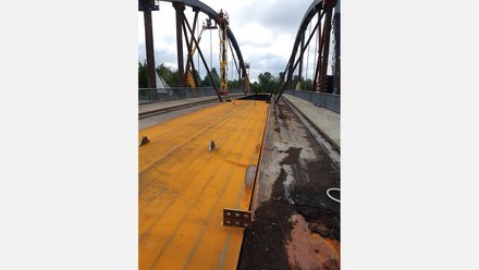 Brückenbau: Gelb‑orange Stahlfahrbahnplatte, mobile Arbeitsbühnen an den Stahlbögen; Monteure arbeiten an der Konstruktion.