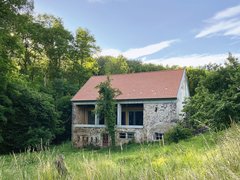 Altes, teils verfallenes Haus mit rotem Ziegeldach inmitten einer hohen Wiese und umgeben von Bäumen unter blauem Himmel