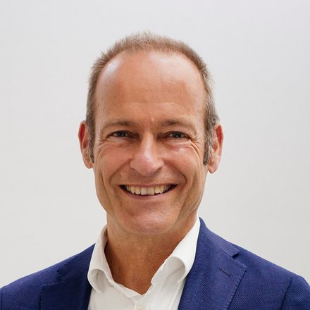Porträtfoto eines Mannes mittleren Alters mit lichtem braunem Haar in einem hellen Büroumfeld. Er trägt einen dunkelblauen Blazer über einem weißen Hemd und lächelt direkt in die Kamera. Im Hintergrund sind verschwommen ein weißer Schreibtisch, ein roter Stuhl und Büromöbel sichtbar.
