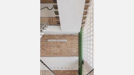 Treppenhaus mit Backsteinwänden  Ein vertikaler Blick in ein Treppenhaus. Sichtbare Betontreppen verlaufen entlang der Wände, die aus rötlichem Backstein bestehen. Große Glasbausteine lassen diffuses Tageslicht in den Raum. Eine grün gestrichene Rohrleitung verläuft senkrecht durch das Bild. Das Treppenhaus verbindet die Geschosse und zeigt den Materialmix aus Beton, Ziegel und Glas. Es vermittelt Orientierung und die robuste, funktionale Bauweise des Bestands.