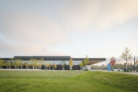 Modernes Gebäude mit verglaster Fassade, umgeben von Rasenflächen und jungen Bäumen unter warmem Abendhimmel.