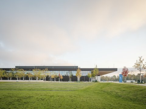 Modernes Gebäude mit verglaster Fassade, umgeben von Rasenflächen und jungen Bäumen unter warmem Abendhimmel.