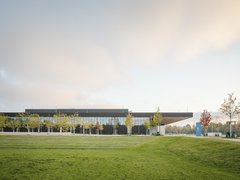 Modernes Gebäude mit verglaster Fassade, umgeben von Rasenflächen und jungen Bäumen unter warmem Abendhimmel.