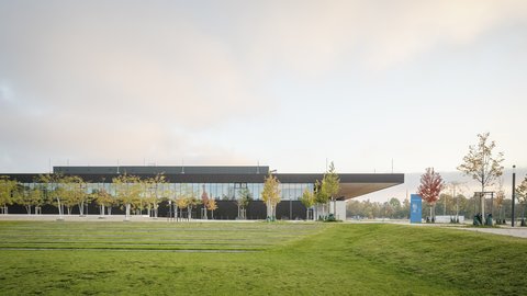 Modernes Gebäude mit verglaster Fassade, umgeben von Rasenflächen und jungen Bäumen unter warmem Abendhimmel.
