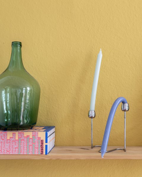 „Ein Holzregal vor einer senfgelben Wand, bestückt mit einer großen grünen Glasflasche auf einer Schachtel, einer Kerze, modernen Deko-Objekten und einer Zimmerpflanze.“