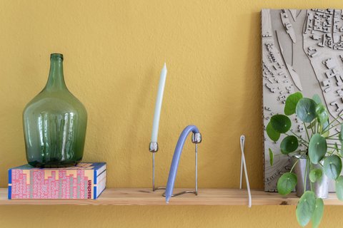 „Ein Holzregal vor einer senfgelben Wand, bestückt mit einer großen grünen Glasflasche auf einer Schachtel, einer Kerze, modernen Deko-Objekten und einer Zimmerpflanze.“