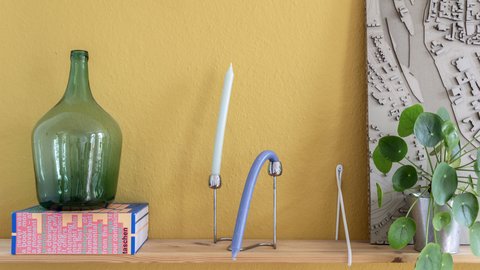„Ein Holzregal vor einer senfgelben Wand, bestückt mit einer großen grünen Glasflasche auf einer Schachtel, einer Kerze, modernen Deko-Objekten und einer Zimmerpflanze.“