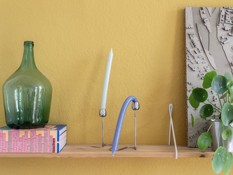 „Ein Holzregal vor einer senfgelben Wand, bestückt mit einer großen grünen Glasflasche auf einer Schachtel, einer Kerze, modernen Deko-Objekten und einer Zimmerpflanze.“