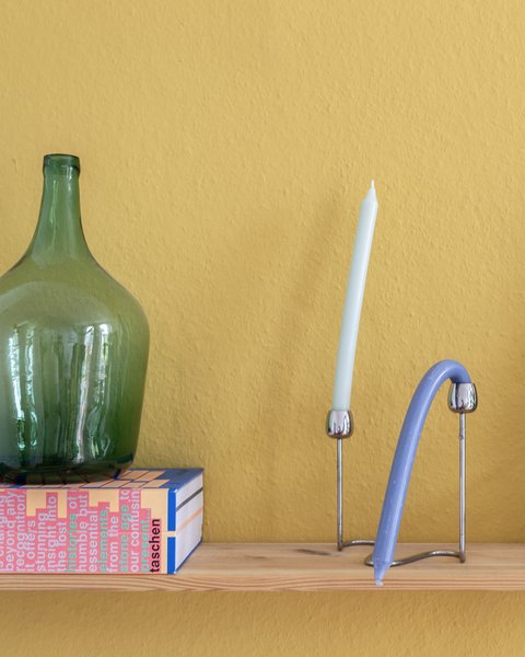„Ein Holzregal vor einer senfgelben Wand, bestückt mit einer großen grünen Glasflasche auf einer Schachtel, einer Kerze, modernen Deko-Objekten und einer Zimmerpflanze.“