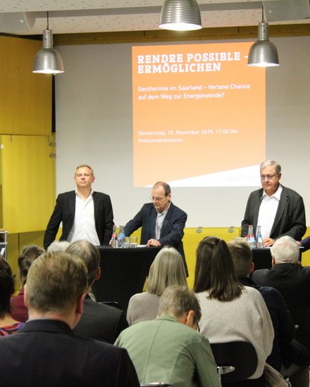 Podiumsdiskussion zur Geothermie  Ein Veranstaltungsraum mit Publikum im Vordergrund und einem Podium im Hintergrund. Fünf Männer stehen auf der Bühne: einer am Rednerpult, vier an Stehtischen mit Wasserflaschen. Hinter ihnen ist eine Präsentationsfolie mit dem Titel „Rendre possible – Ermöglichen. Geothermie im Saarland – Vertane Chance auf dem Weg zur Energiewende?“ sowie Datum und Uhrzeit der Veranstaltung. Das Bild zeigt eine öffentliche Podiumsdiskussion zu Energiepolitik und Geothermie. Es steht für fachlichen Austausch, politische Debatte und gesellschaftliche Auseinandersetzung mit der Energiewende. 