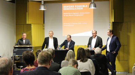 Podiumsdiskussion zur Geothermie  Ein Veranstaltungsraum mit Publikum im Vordergrund und einem Podium im Hintergrund. Fünf Männer stehen auf der Bühne: einer am Rednerpult, vier an Stehtischen mit Wasserflaschen. Hinter ihnen ist eine Präsentationsfolie mit dem Titel „Rendre possible – Ermöglichen. Geothermie im Saarland – Vertane Chance auf dem Weg zur Energiewende?“ sowie Datum und Uhrzeit der Veranstaltung. Das Bild zeigt eine öffentliche Podiumsdiskussion zu Energiepolitik und Geothermie. Es steht für fachlichen Austausch, politische Debatte und gesellschaftliche Auseinandersetzung mit der Energiewende. 