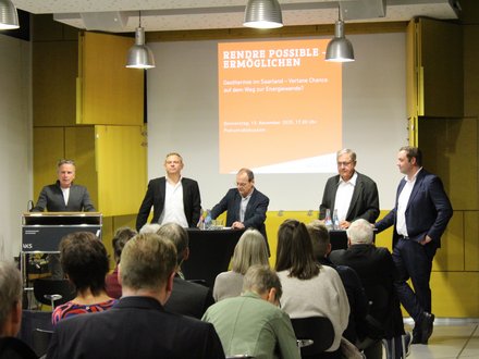 Podiumsdiskussion zur Geothermie  Ein Veranstaltungsraum mit Publikum im Vordergrund und einem Podium im Hintergrund. Fünf Männer stehen auf der Bühne: einer am Rednerpult, vier an Stehtischen mit Wasserflaschen. Hinter ihnen ist eine Präsentationsfolie mit dem Titel „Rendre possible – Ermöglichen. Geothermie im Saarland – Vertane Chance auf dem Weg zur Energiewende?“ sowie Datum und Uhrzeit der Veranstaltung. Das Bild zeigt eine öffentliche Podiumsdiskussion zu Energiepolitik und Geothermie. Es steht für fachlichen Austausch, politische Debatte und gesellschaftliche Auseinandersetzung mit der Energiewende. 