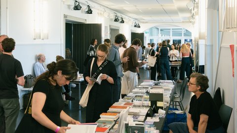 Innenansicht einer belebten Buchmesse oder Ausstellung in einem hellen, modernen Flur. Besucher stehen an langen Tischen, die dicht mit Architekturmagazinen und Büchern gefüllt sind, blättern in den Publikationen und tauschen sich aus.