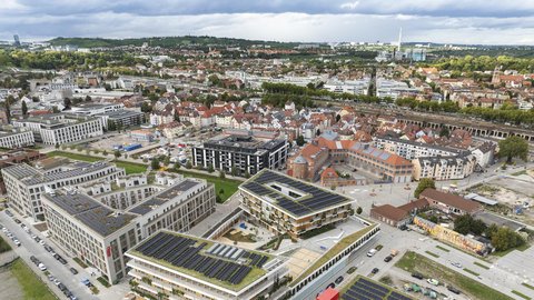 Luftaufnahme eines Stadtteils mit mehreren modernen Gebäuden, viele davon mit Solarpanelen auf den Dächern, umgeben von Straßen und weiteren Wohnhäusern