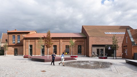 Backsteingebäude mit mehreren Giebeln und einem kleinen Springbrunnen auf gepflastertem Platz davor, zwei Personen gehen am Brunnen vorbei