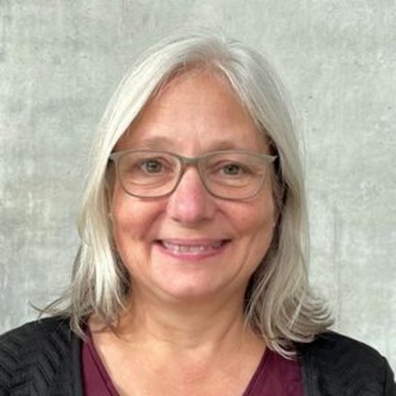 Porträt einer lächelnden Person mit schulterlangem grauem Haar und Brille vor neutral grauer Wand.