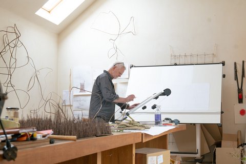 Älterer Mann mit grauem Hemd arbeitet an einem großen Zeichenbrett in einem Atelier mit Skulpturen und Kunstmaterialien