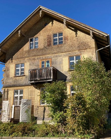 Ein freistehendes historisches Gebäude mit Satteldach, verputzter Fassade und kleinem Holzbalkon steht in einer ländlich geprägten Umgebung mit Bäumen und Sträuchern, wodurch das Bild ein erhaltenes oder umgenutztes Schul- oder Wohnhaus zeigt und für den behutsamen Umgang mit regionaler Baukultur, Bestandspflege und lokaler Identität steht.