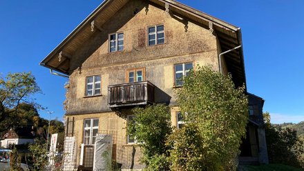 Ein freistehendes historisches Gebäude mit Satteldach, verputzter Fassade und kleinem Holzbalkon steht in einer ländlich geprägten Umgebung mit Bäumen und Sträuchern, wodurch das Bild ein erhaltenes oder umgenutztes Schul- oder Wohnhaus zeigt und für den behutsamen Umgang mit regionaler Baukultur, Bestandspflege und lokaler Identität steht.