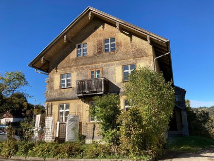 Ein freistehendes historisches Gebäude mit Satteldach, verputzter Fassade und kleinem Holzbalkon steht in einer ländlich geprägten Umgebung mit Bäumen und Sträuchern, wodurch das Bild ein erhaltenes oder umgenutztes Schul- oder Wohnhaus zeigt und für den behutsamen Umgang mit regionaler Baukultur, Bestandspflege und lokaler Identität steht.