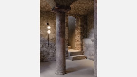"Unterirdischer Raum mit rauen Steinmauern. Eine dicke zylindrische Säule mit Kapitäll steht zentral. Links an der Wand zwei runde Wandleuchten an schwarzem Rohr. Zwei breite Betonstufen führen zu einer dunklen Öffnung. Der Boden ist aus hellem Beton."