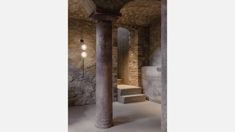 "Unterirdischer Raum mit rauen Steinmauern. Eine dicke zylindrische Säule mit Kapitäll steht zentral. Links an der Wand zwei runde Wandleuchten an schwarzem Rohr. Zwei breite Betonstufen führen zu einer dunklen Öffnung. Der Boden ist aus hellem Beton."