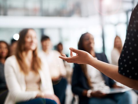 Nahaufnahme einer gestikulierenden Hand einer vortragenden Person; im Hintergrund unscharf ein sitzendes Publikum in einem hellen Seminarraum.