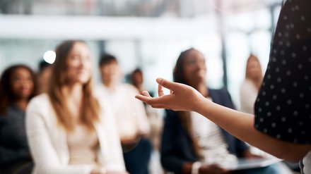 Nahaufnahme einer gestikulierenden Hand einer vortragenden Person; im Hintergrund unscharf ein sitzendes Publikum in einem hellen Seminarraum.