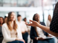 Nahaufnahme einer gestikulierenden Hand einer vortragenden Person; im Hintergrund unscharf ein sitzendes Publikum in einem hellen Seminarraum.